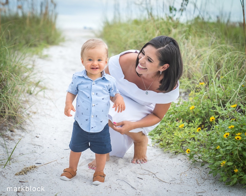 A smiling woman in a white dress kneels on a sandy path next to a happy toddler boy in blue, surrounded by greenery and yellow flowers. The beach is visible in the background—captured beautifully by a Naples beach portrait photographer.