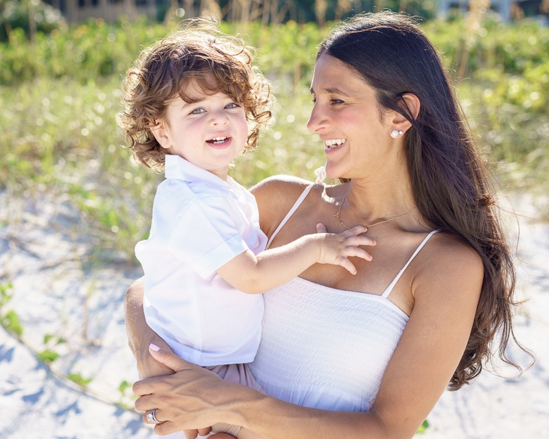 A woman with long dark hair, wearing a white dress, smiles while holding a young child in a white shirt. Captured by a Naples beach portrait photographer, they stand outside in bright sunlight, surrounded by greenery and sand.