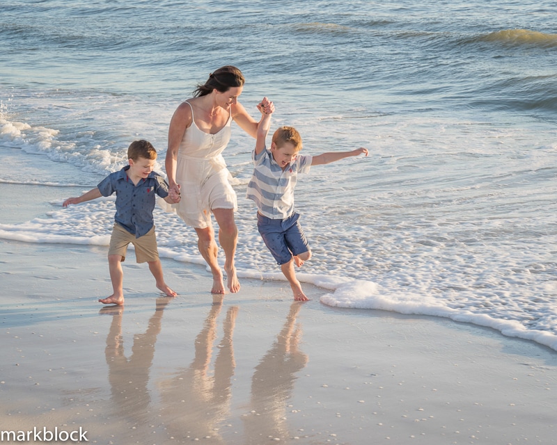 A woman in a white dress holds hands with two young boys as they run and play by the ocean’s edge, gentle waves washing onto the sand—an unforgettable moment captured by a Naples beach portrait photographer.