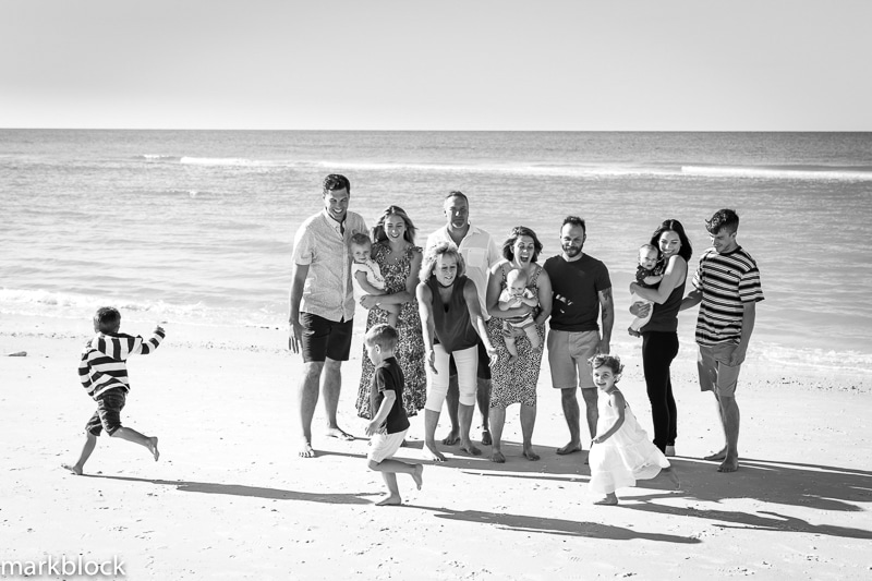 A group of adults and young children pose and play together on a sunny beach, smiling and laughing. The ocean shimmers in the background of this black-and-white photo by a Naples beach portrait photographer.