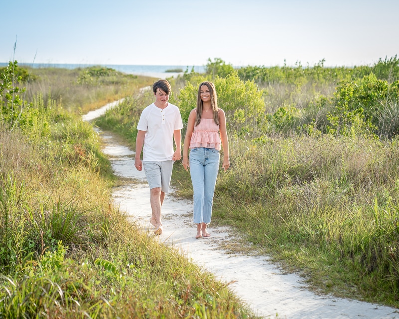 A young man and woman walk barefoot together on a sandy path through grassy dunes, smiling, with greenery around them and the beach visible in the distance under a clear sky—captured by a Naples beach portrait photographer.