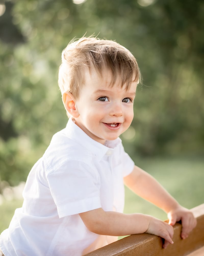 A young boy with light brown hair and a white shirt smiles while holding onto a wooden railing outdoors, captured beautifully by a Naples beach portrait photographer against a blurred green background.