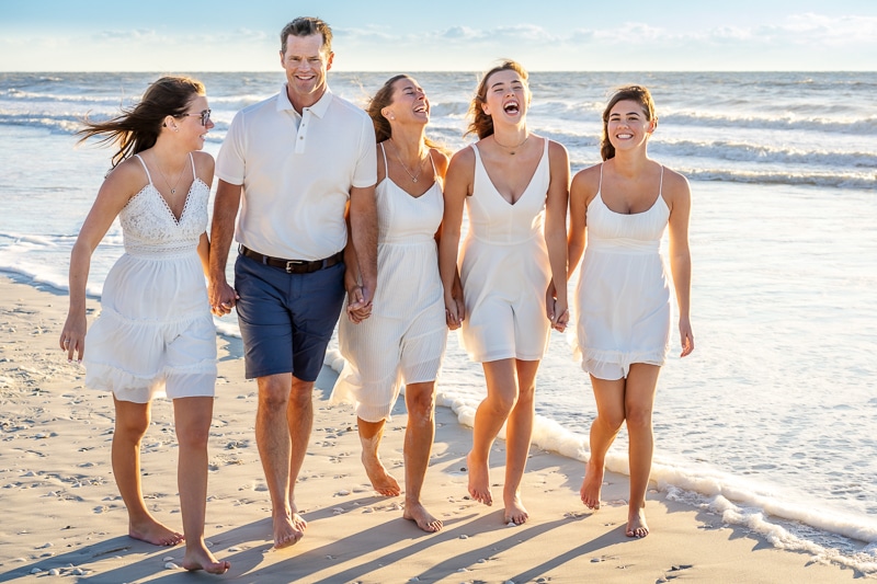 A man and four women in white outfits walk barefoot along the shore, smiling and laughing together beneath a clear sky—captured perfectly by a Naples beach portrait photographer.