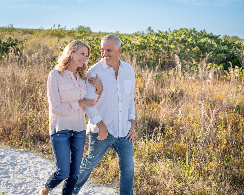 A happy older couple walks arm in arm along a sandy path, smiling at each other, with tall grass and green shrubs behind them under a clear blue sky—captured beautifully by a Naples beach portrait photographer.