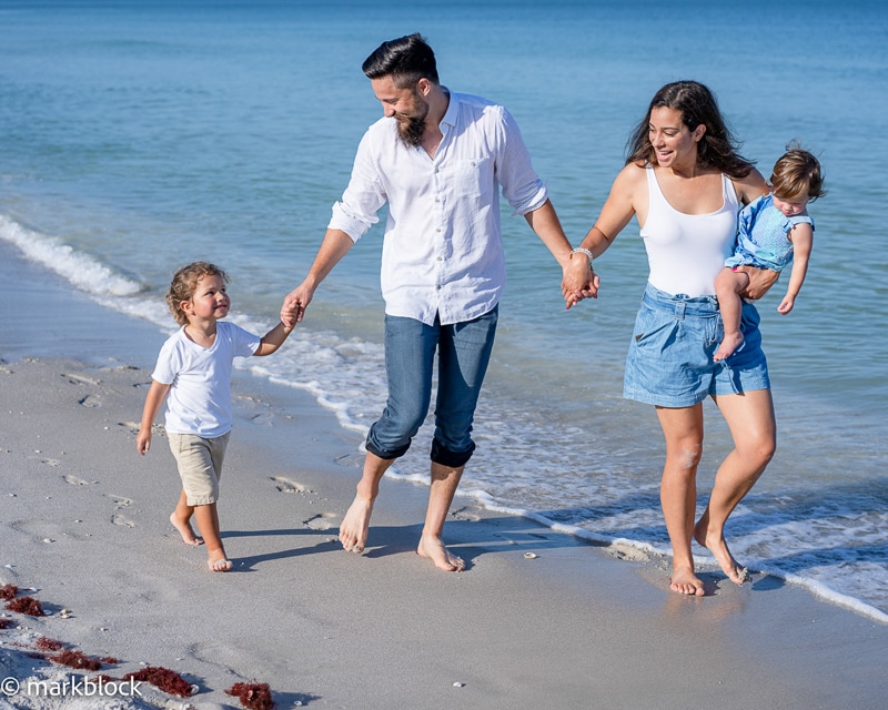 A smiling family of four walks barefoot along the beach, perfectly captured by a Naples beach portrait photographer. The father holds hands with a child while the mother, in a white top and blue skirt, carries a toddler under the clear sky.