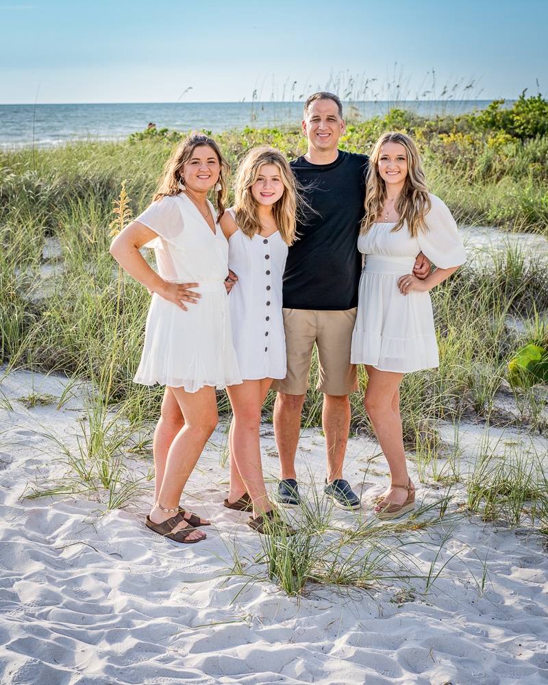 A man and three young women stand together on a sandy beach with sea grass, all smiling. Captured by a Naples beach portrait photographer, the women wear white dresses and the man wears a black shirt and khaki shorts with the ocean behind them.