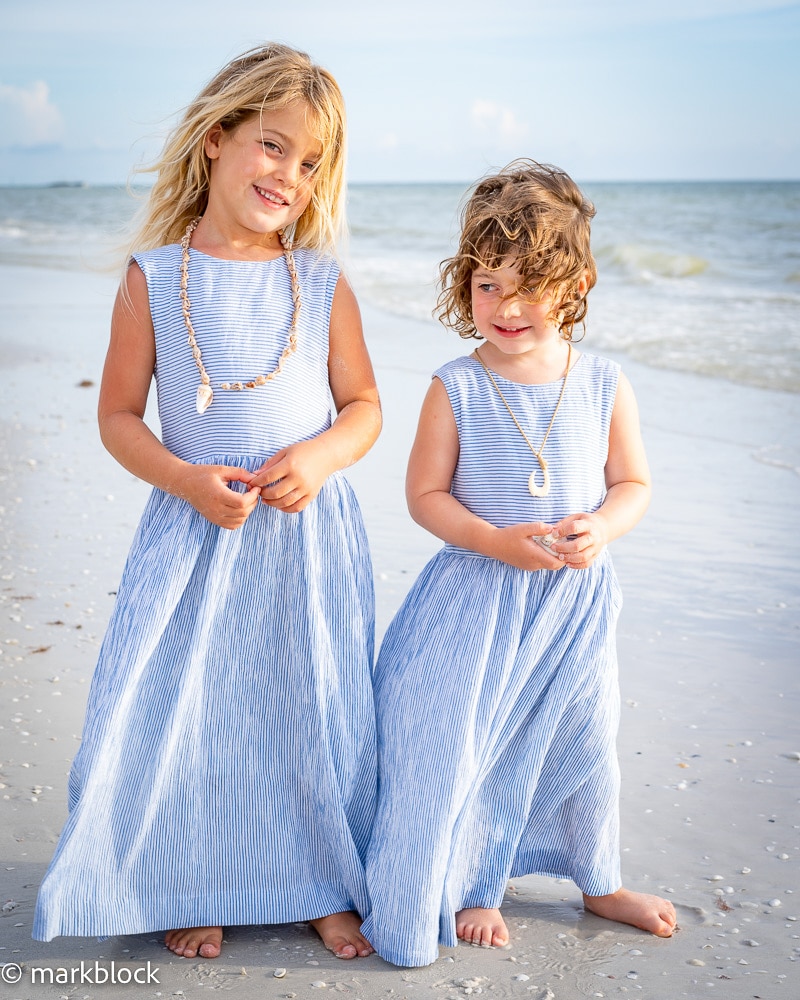 Two young children in matching blue and white striped dresses stand barefoot on a sandy beach, smiling brightly as the wind blows their hair—an ideal scene captured by a Naples beach portrait photographer, with the ocean as their backdrop.