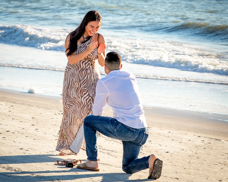 A man kneels on one knee proposing to a woman on a sandy beach near the ocean, captured by a Naples beach portrait photographer. The woman, in a patterned dress, looks surprised and happy as waves roll in behind them.