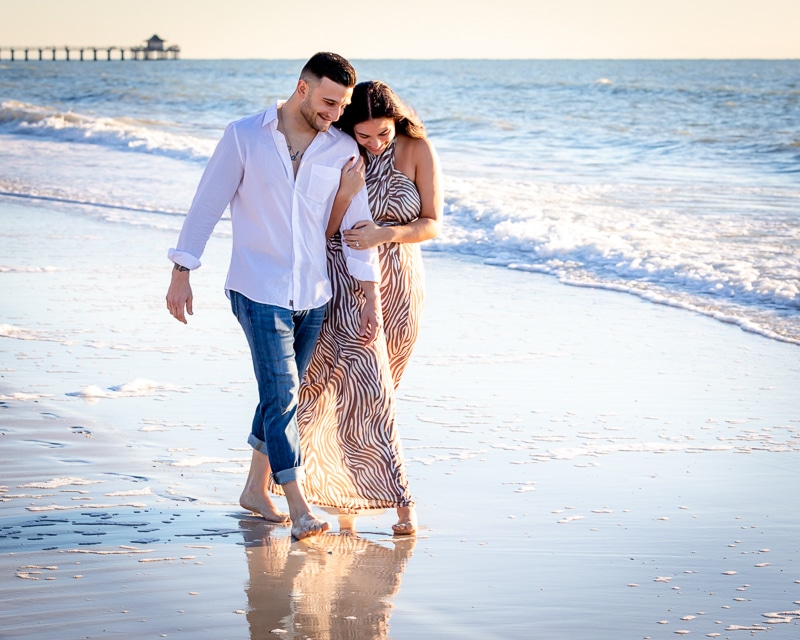 A couple walks barefoot along the shoreline, smiling and holding each other. Captured by a Naples beach portrait photographer, the woman wears a flowing striped dress while the man sports rolled-up jeans and a white shirt, with the ocean and distant pier behind them.