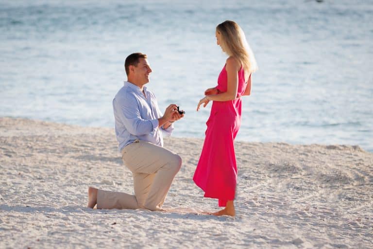 A man kneels on a sandy beach holding a ring box, proposing to a woman in a bright pink dress near the ocean—captured perfectly by a Naples beach portrait photographer.