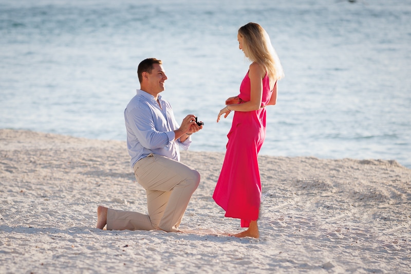 A man kneels on a sandy beach holding a ring box, proposing to a woman in a bright pink dress near the ocean—captured perfectly by a Naples beach portrait photographer.