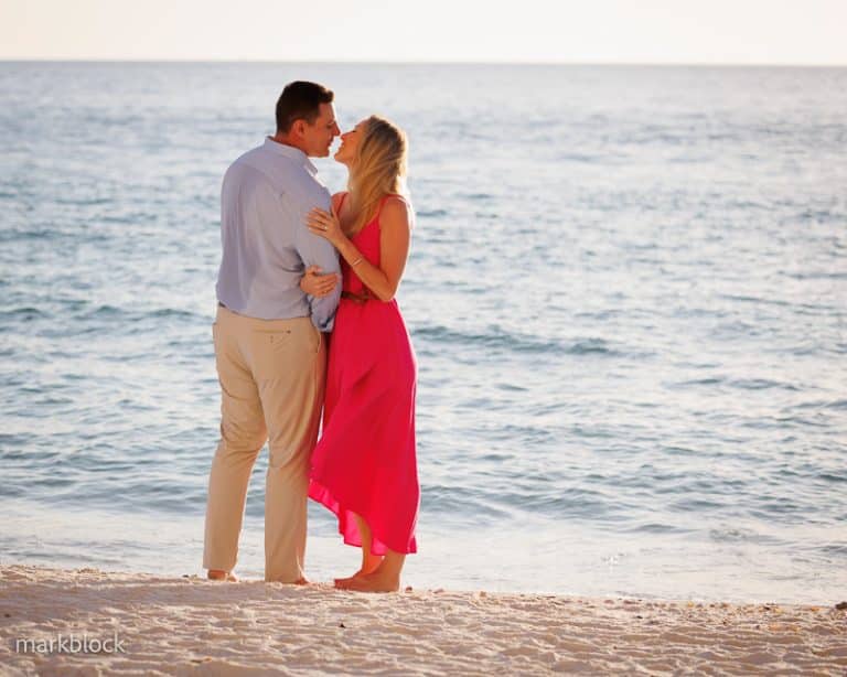 A couple stands on a sandy Naples FL beach at sunset, embracing and about to kiss. Captured by a Naples beach portrait photographer, the man wears light pants and a blue shirt, while the woman glows in a bright pink dress with ocean waves in the background.