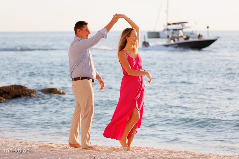 A man and woman dance barefoot on a sandy beach at sunset. The woman wears a bright pink dress and the man wears a light blue shirt and beige pants—a perfect scene for any Naples beach portrait photographer. A boat is visible on the ocean in the background.
