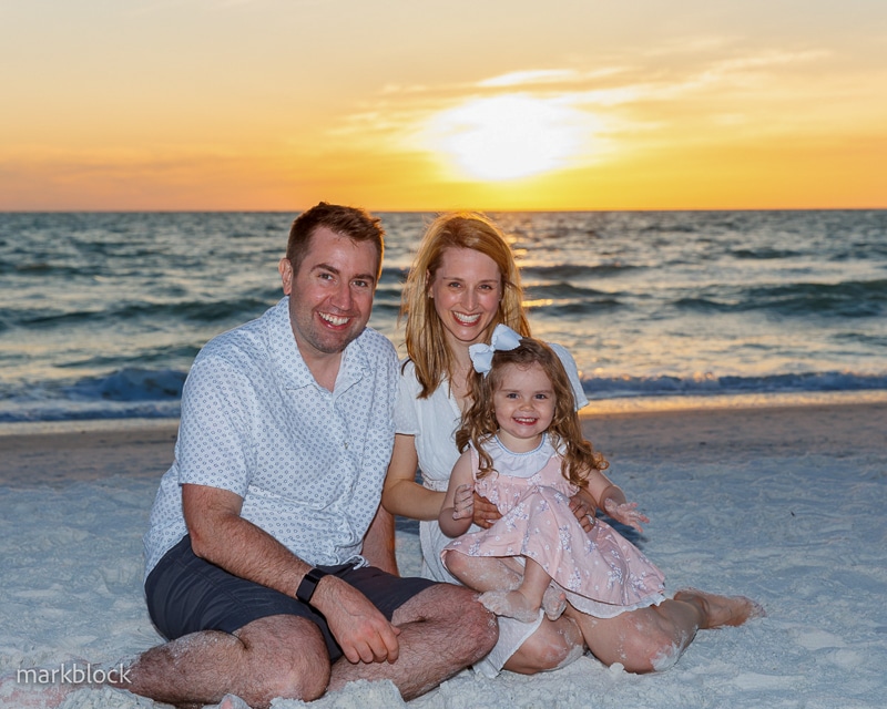 A smiling family of three—a man, a woman, and a young girl in a pink dress—sits on sandy beach at sunset with waves in the background, beautifully captured by a Naples beach portrait photographer.