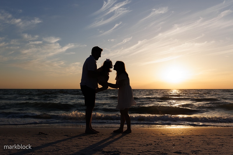 A family of three stands on the beach at sunset, silhouetted against the colorful sky and ocean waves—a perfect moment captured by a Naples beach portrait photographer.