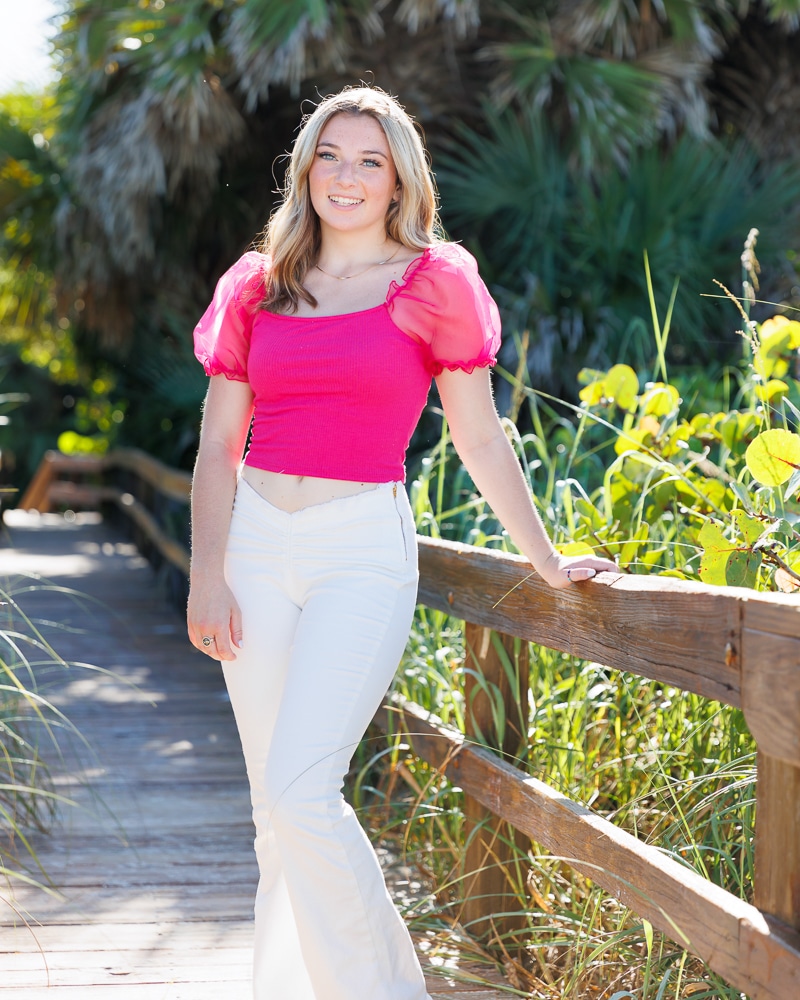 A young woman with long blonde hair, wearing a bright pink top and white pants, stands smiling on a wooden boardwalk surrounded by lush green plants and sunlight, captured perfectly by a Naples beach portrait photographer.