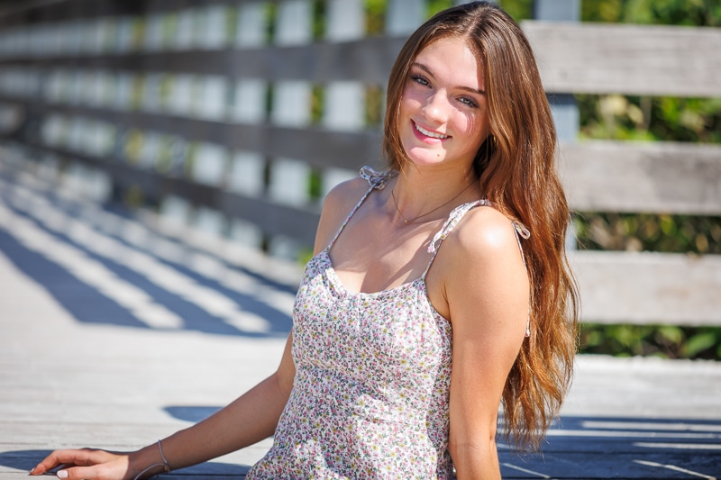 A young woman with long brown hair, wearing a floral dress, sits on a wooden walkway outdoors, smiling at the camera in bright sunlight with green foliage behind her—a perfect moment for a Naples beach portrait photographer.