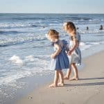 Two young girls in blue dresses stand barefoot at the edge of the Naples FL beach, watching the waves on a sandy beach under a clear sky—an ideal moment for a Naples beach portrait photographer to capture.