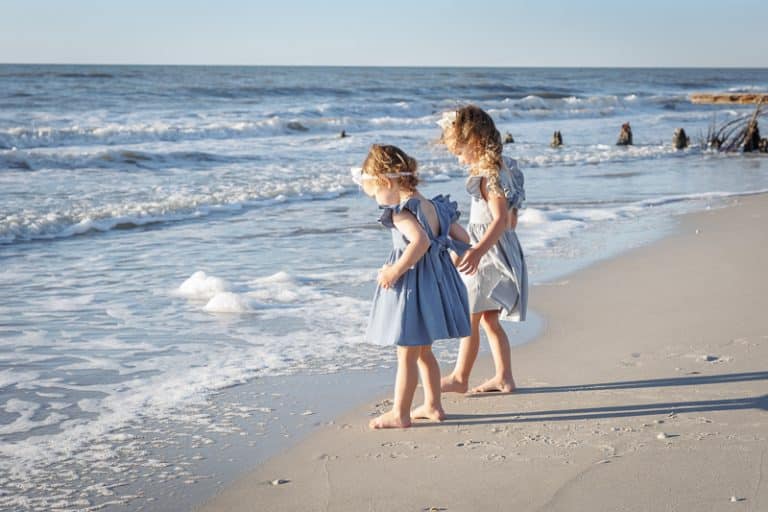 Two young girls in blue dresses stand barefoot at the edge of the Naples FL beach, watching the waves on a sandy beach under a clear sky—an ideal moment for a Naples beach portrait photographer to capture.