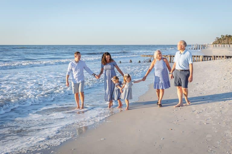 A family of six, including two young girls, walks hand-in-hand along a sunny Naples beach. Barefoot and dressed in summery clothes, they enjoy the waves—captured perfectly by a Naples beach portrait photographer.