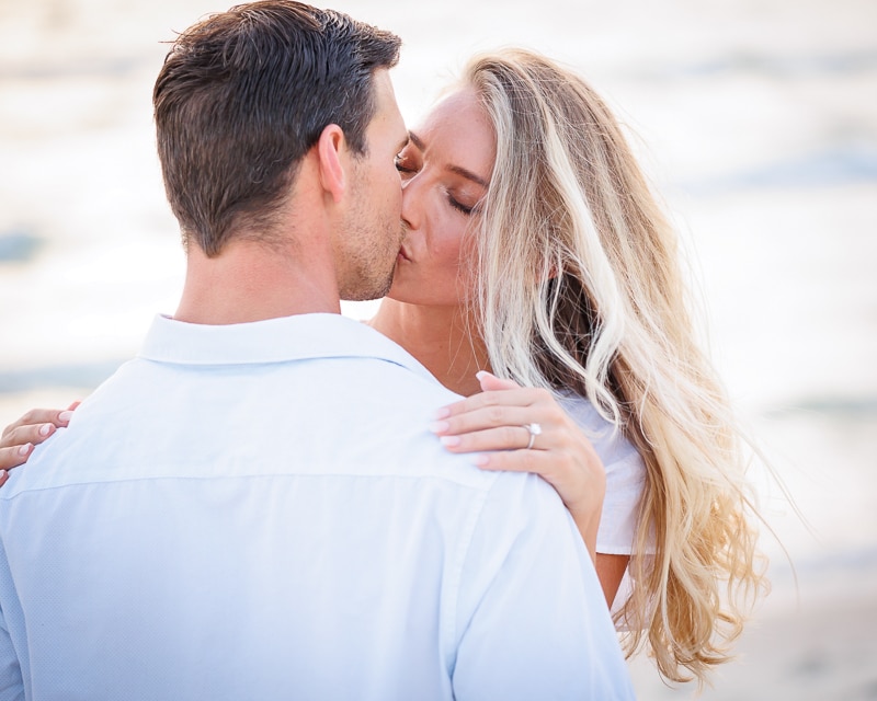 A couple shares a kiss on the beach. The woman, with long blonde hair, has her arms around the man's neck and displays an engagement ring. Captured by a Naples beach portrait photographer, both are dressed in white against a softly blurred background.