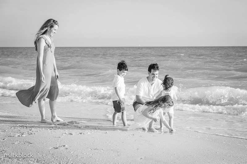 A family of five, captured by a Naples beach portrait photographer, enjoys time together at the shoreline. The woman stands on the sand while the man and children play near the water in this striking black and white scene.