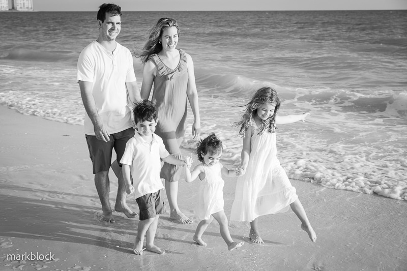 A smiling family of five, including two adults and three young children, walks barefoot along the shore. Captured by a Naples beach portrait photographer, the kids play joyfully as gentle waves roll in under clear skies.