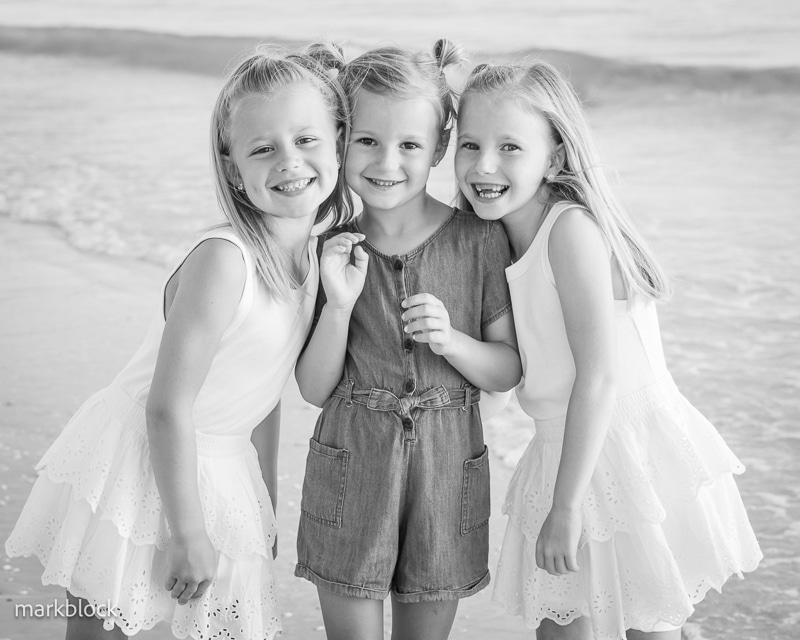Three young girls smile and pose together on a beach. Captured by a Naples beach portrait photographer, two wear white dresses while the center girl dons a short-sleeved romper. The ocean and shoreline form the black-and-white backdrop.