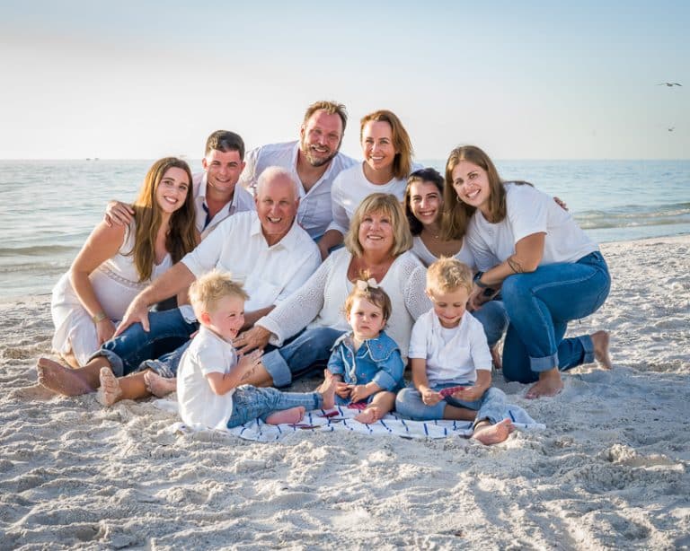 A multi-generational family, all in white and denim outfits, smiles at the camera while sitting on a sandy beach. The ocean and sky frame the scene—captured perfectly by a Naples beach portrait photographer. Three young children sit in front.