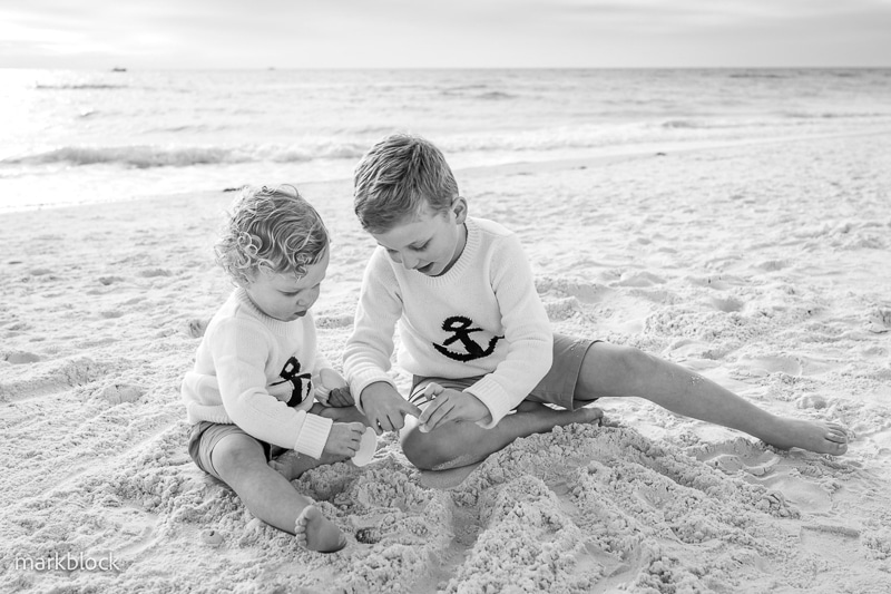 Two young children in matching anchor sweaters sit on a sandy beach, playing together. Captured by a Naples beach portrait photographer, the black and white photo shows them digging in the sand with the ocean and sky in the background.