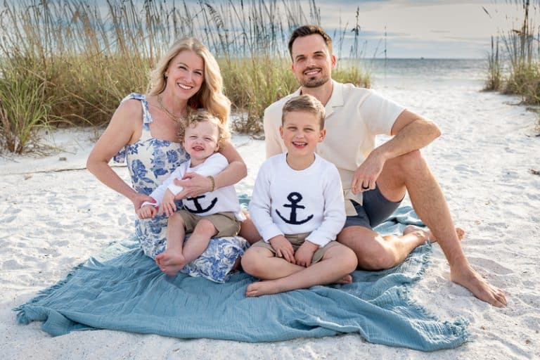 A family of four sits on a blue blanket on a sandy beach. Captured by a Naples beach portrait photographer, the mother holds a young child while the father sits with an older one, all smiling in light, summery clothes with ocean and grass behind them.