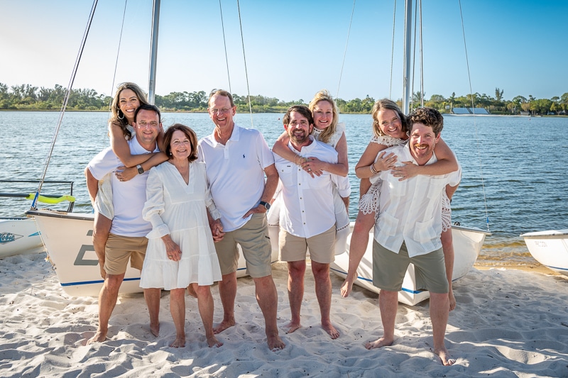 A group of eight smiling adults pose on a sandy beach in front of sailboats; three women are playfully piggyback on men, all dressed in light casual summer clothing, captured by a Naples beach portrait photographer.
