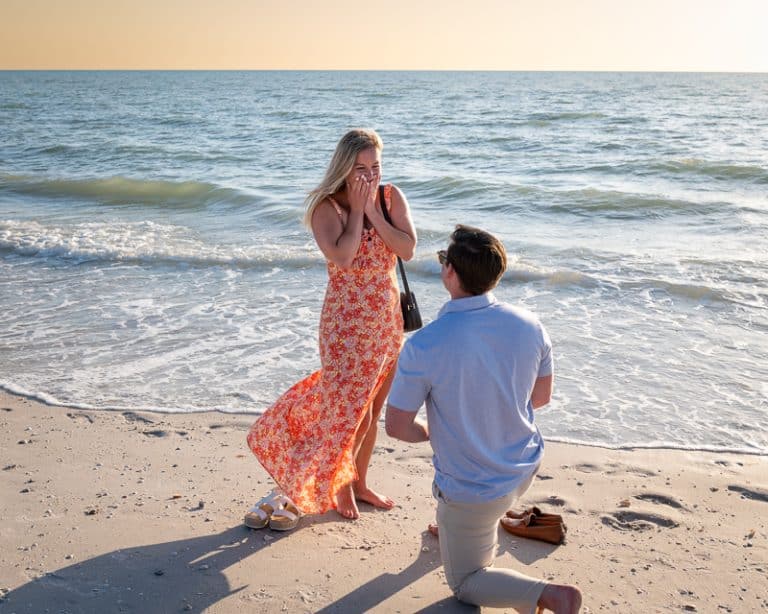 A person kneels on a sandy beach proposing to a surprised woman in a flowing orange floral dress, with gentle waves and a calm sea at sunset—captured perfectly by a Naples beach portrait photographer.