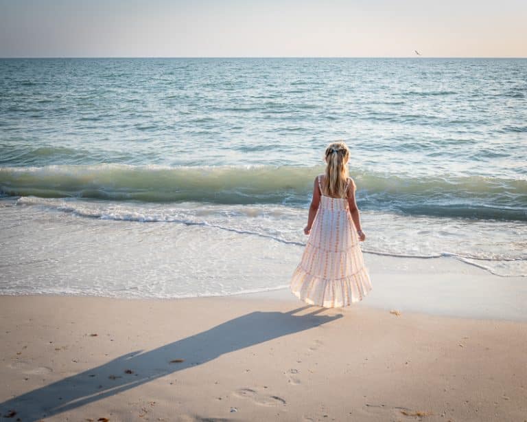 A young girl in a long, light dress stands on a sandy beach, facing the ocean with gentle waves under a clear sky—her shadow stretching behind her, captured perfectly by a Naples beach portrait photographer.