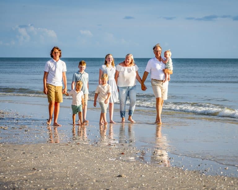 A family of seven, including two adults and five children, walks barefoot along the ocean, all smiling and holding hands under a bright sky—perfect for a Naples beach portrait photographer to capture this joyful moment.