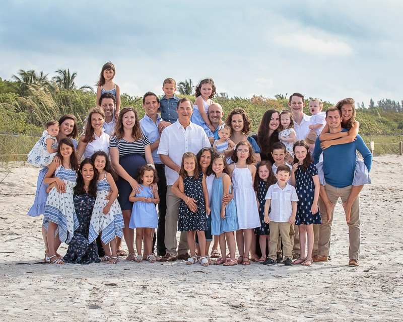 A large multigenerational family poses together on a sandy beach, smiling at the camera. Captured by a Naples beach portrait photographer, adults, children, and toddlers wear coordinated blue and white outfits with greenery and a cloudy sky behind them.