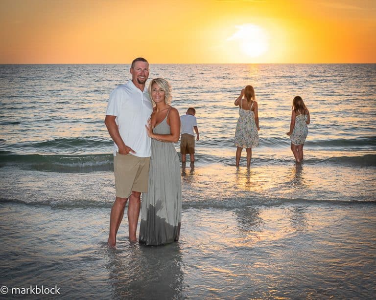 A couple stands smiling on the beach at sunset with waves at their feet, while three children play behind them in the shallow water—captured beautifully by a Naples beach portrait photographer under a golden, tranquil sky.