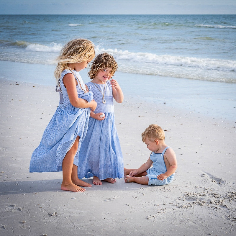 Two young girls in blue dresses stand on a sandy beach near the water, while a toddler boy in blue overalls plays in the sand—captured beautifully by a Naples beach portrait photographer as waves roll under a cloudy sky.