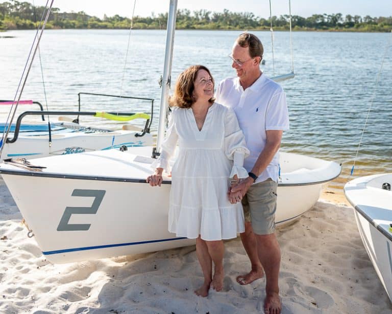 A smiling couple stands barefoot on sandy ground beside a small sailboat marked with the number 2, holding hands. Captured by a Naples beach portrait photographer, the water and more boats are visible in the background on a sunny day.