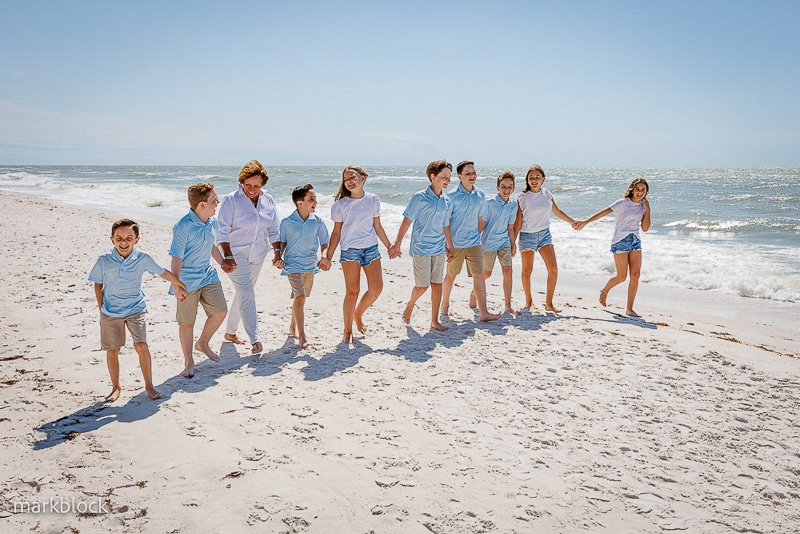 A group of children and teens in blue and white clothes hold hands, smiling and laughing as they walk along a sunny beach, captured perfectly by a Naples beach portrait photographer with the ocean in the background.