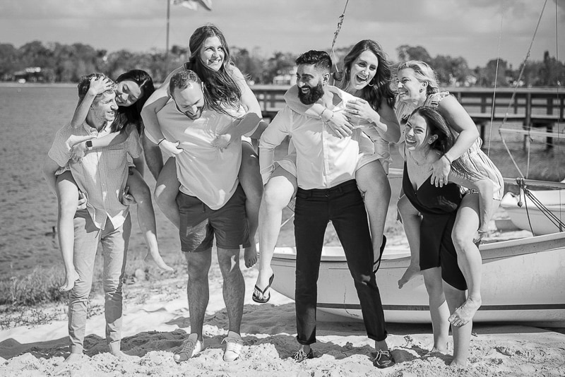 A group of eight people on a sandy beach, four women riding piggyback on four men. Captured by a Naples beach portrait photographer, they are all laughing and smiling, with boats and water in the background. The image is in black and white.