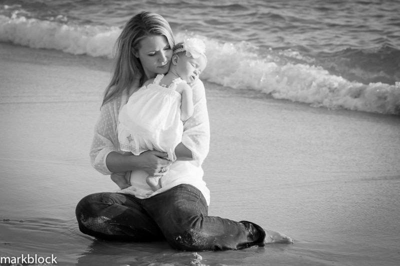 A woman kneels on the beach, holding a sleeping baby in her arms. Both in light clothing, they are framed by gentle waves in this black and white image captured by a Naples beach portrait photographer.