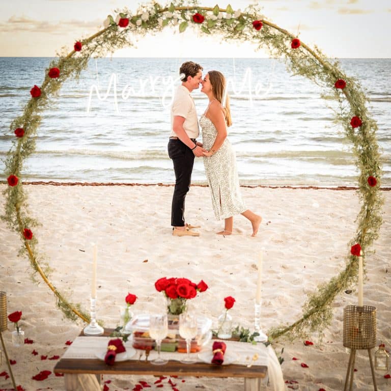 A couple stands barefoot on a beach inside a circular floral arch, holding hands and looking at each other. Captured by a Naples beach portrait photographer, the scene includes a decorated table with candles and red roses, against the ocean backdrop.