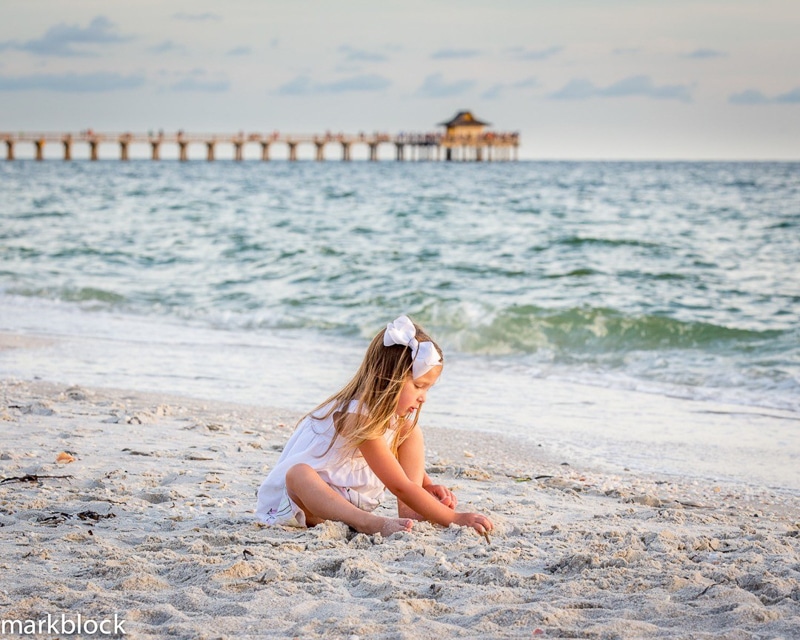 A young girl in a white dress and bow sits on a sandy beach, playing with sand. The ocean waves approach the shore while a pier extends into the water under a partly cloudy sky—an ideal moment for any Naples beach portrait photographer.