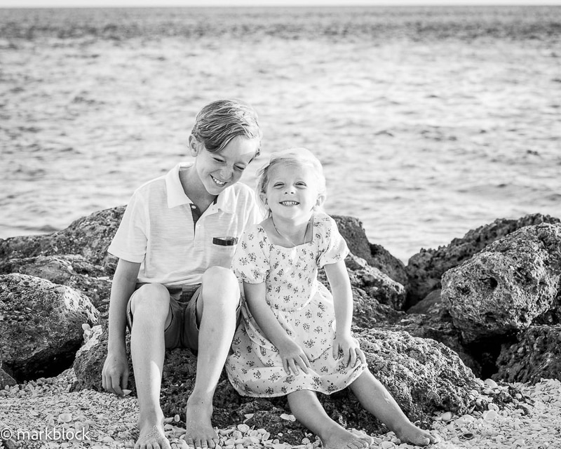 Two young children, a boy and a girl, sit barefoot on rocks by the seaside, smiling happily at the camera. Captured by a Naples beach portrait photographer, this black and white image features the ocean in the background.