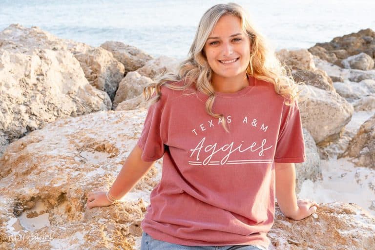 A young woman with long blonde hair sits on rocks at the beach, smiling and wearing a maroon Texas A&M Aggies T-shirt. Captured by a Naples beach portrait photographer, the ocean and more rocks are visible in the background.