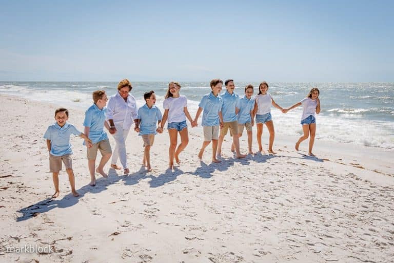 A group of ten children and teens, dressed in light blue shirts, white tops, and khaki shorts, hold hands and walk along a sunny beach with waves in the background—captured by a Naples beach portrait photographer.