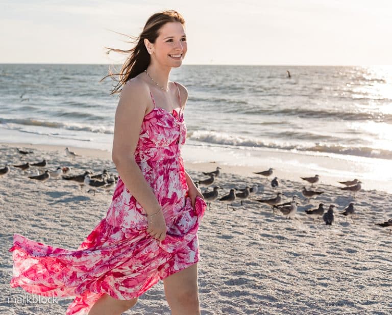 A woman in a pink floral dress walks along a sandy beach with the ocean and a flock of birds behind her on a sunny day. Her hair blows in the breeze, and she smiles—an ideal moment for any Naples beach portrait photographer.