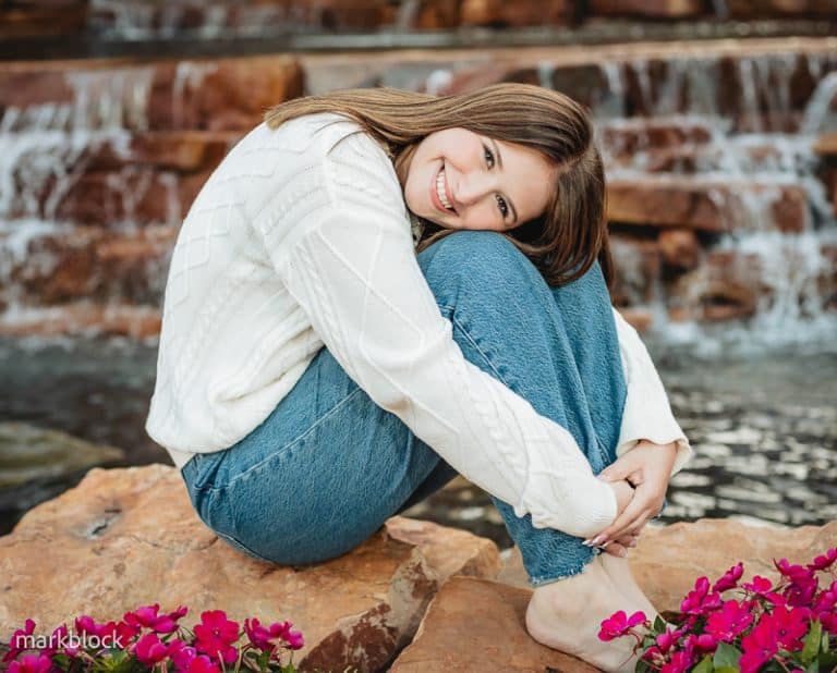 A young woman in a white sweater and blue jeans sits barefoot on a rock, hugging her knees and smiling, as though captured by a Naples beach portrait photographer, with a cascading waterfall and pink flowers in the background.