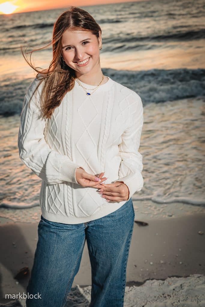 A young woman with long brown hair smiles at the camera while standing on a beach at sunset. Captured by a Naples beach portrait photographer, she wears a white sweater and blue jeans, with ocean waves and a colorful sky in the background.